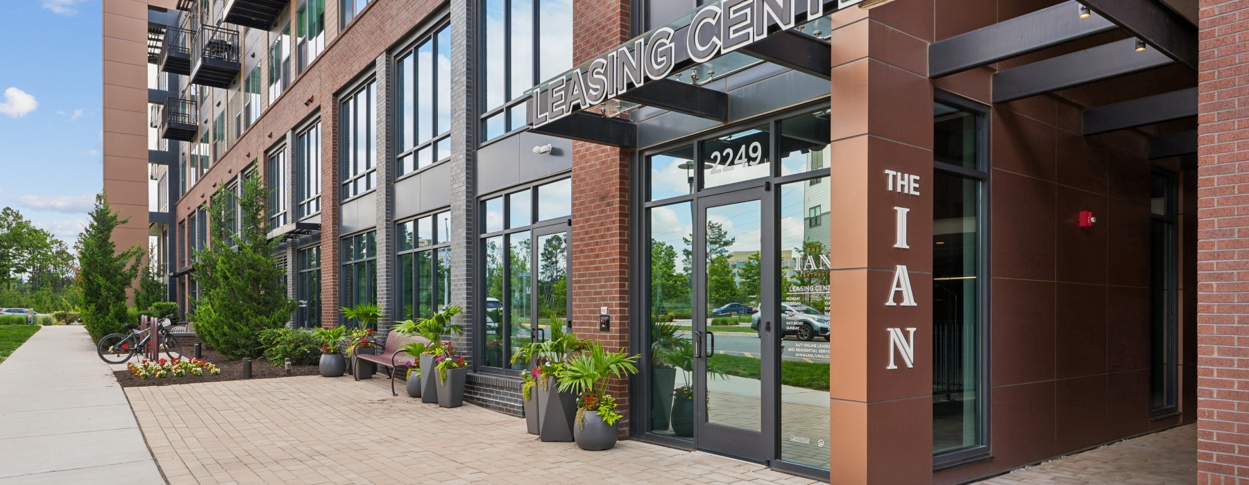 Leasing center entrance at The Ian apartments in Herndon, VA, with a modern brick exterior, glass doors, and a landscaped paver plaza
